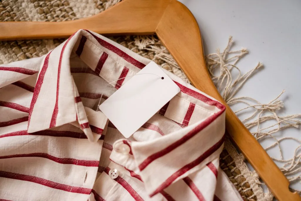 From above of casual shirt with red stripes and blank paper label placed on wooden hanger on rug